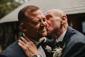 Two people in dark suits share an emotional moment with baby's breath boutonnieres at an outdoor wedding.
