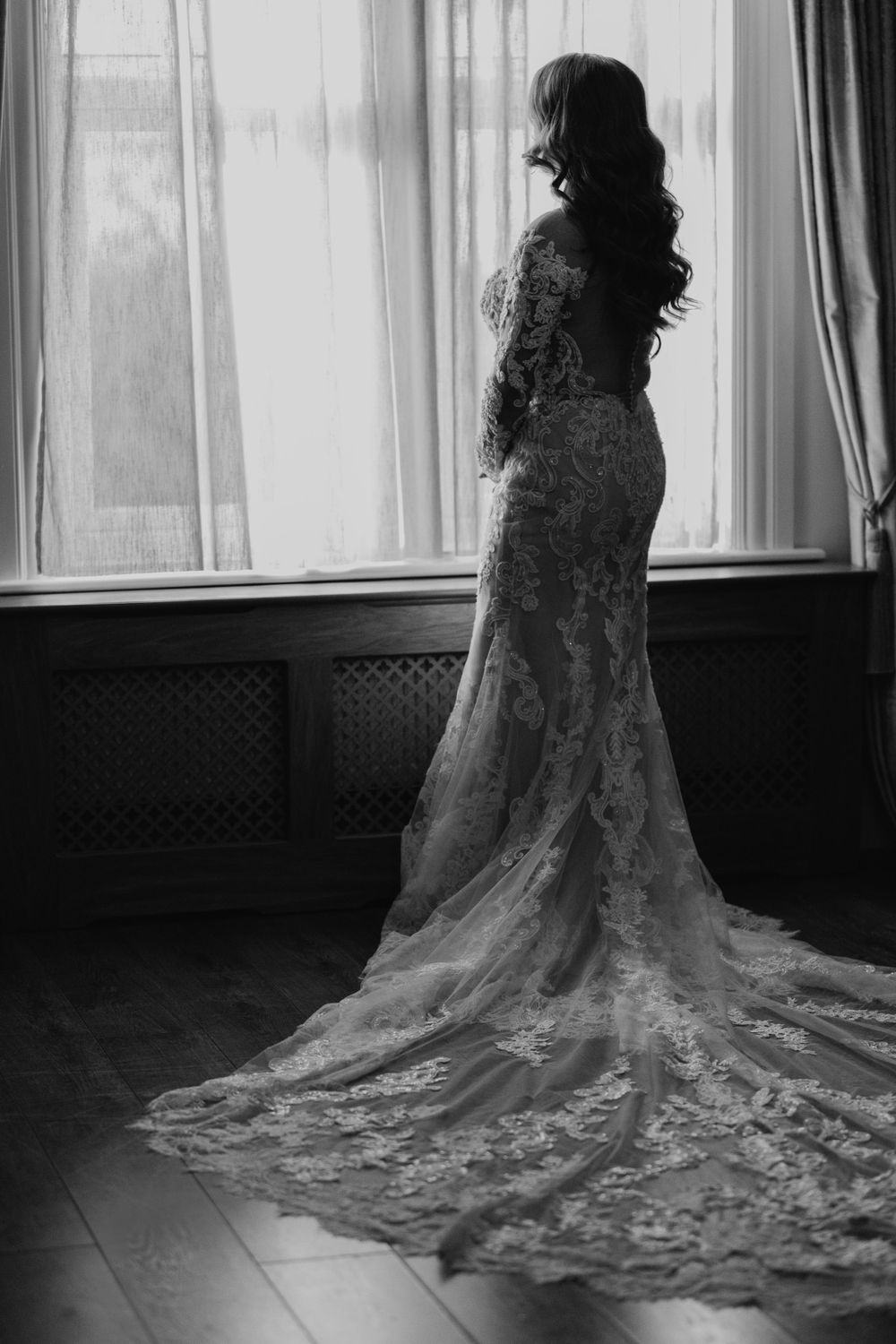 Black and white image of bride in lace gown with long train standing by window.