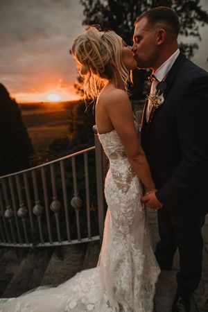 Wedding couple share an intimate sunset moment on a balcony with dramatic orange sky in the background.