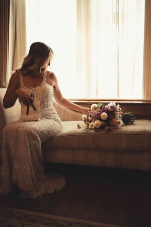 Dreamy vintage-style portrait of someone in a white lace dress sitting by a sunlit window with flowers beside them.