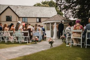 A small dog walks down the aisle during an outdoor wedding ceremony with guests seated in white chairs on the lawn.