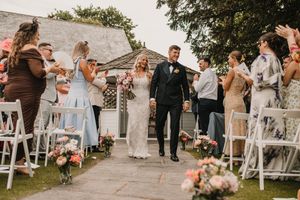 Wedding party celebrating as newlyweds walk down the outdoor aisle lined with floral arrangements and white chairs.
