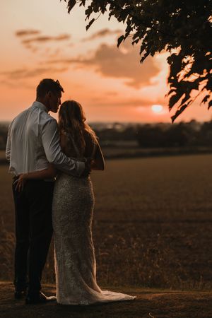 A couple embraces while silhouetted against a warm orange sunset in a rural field.