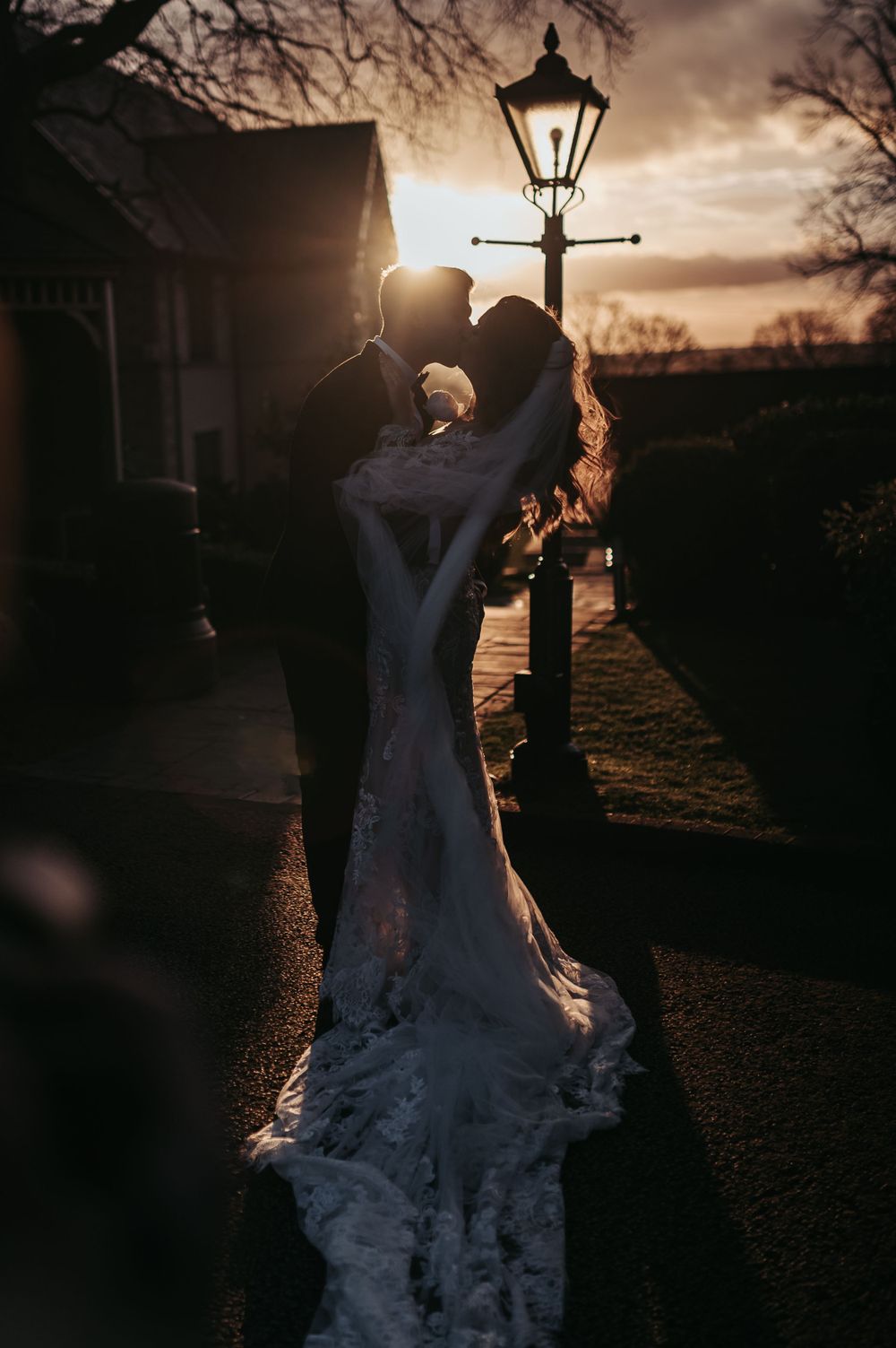 Romantic silhouette of couple embracing near vintage lamppost at sunset in wedding attire.