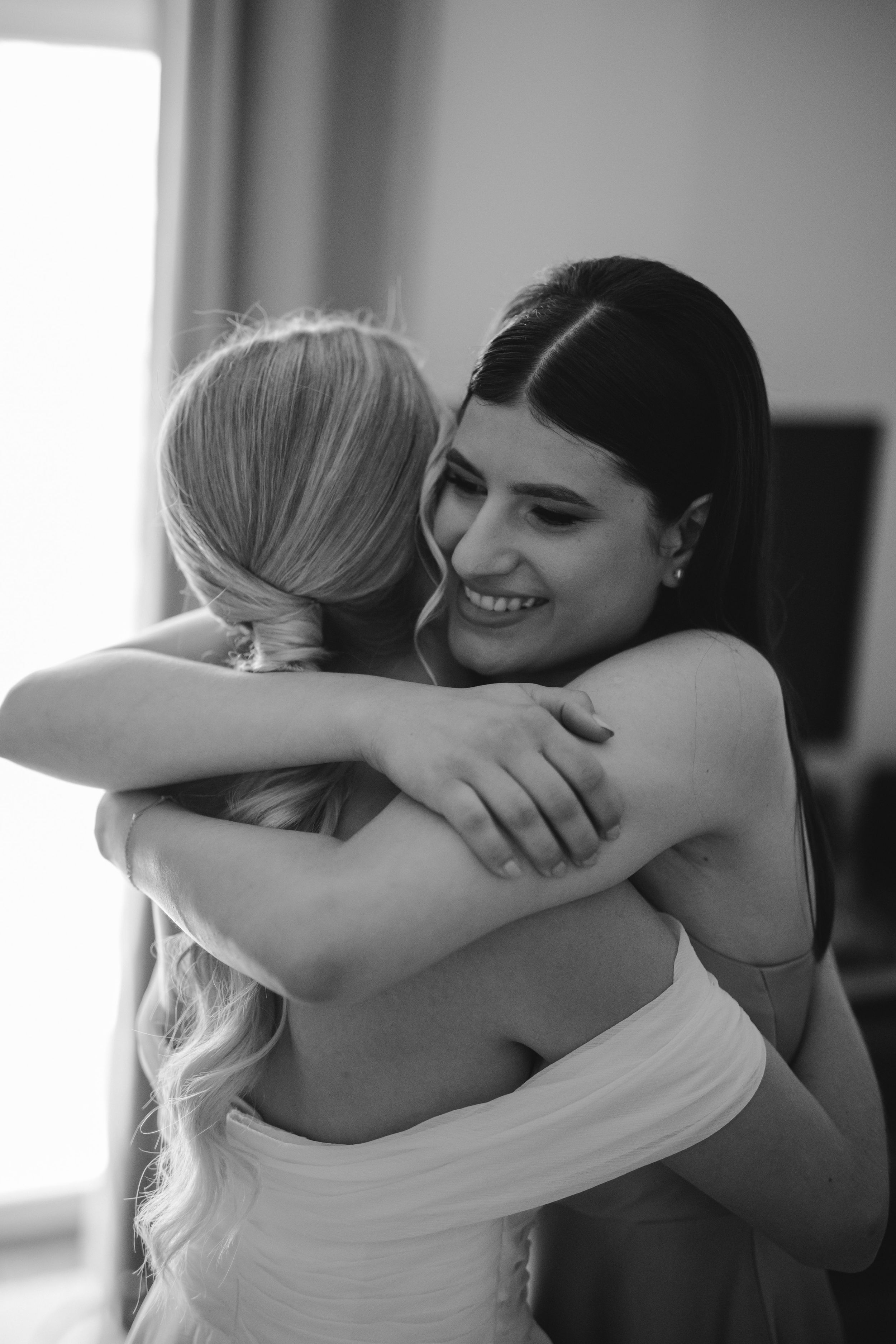 Two women share an emotional embrace at a wedding, one in a white dress and one in dark attire.