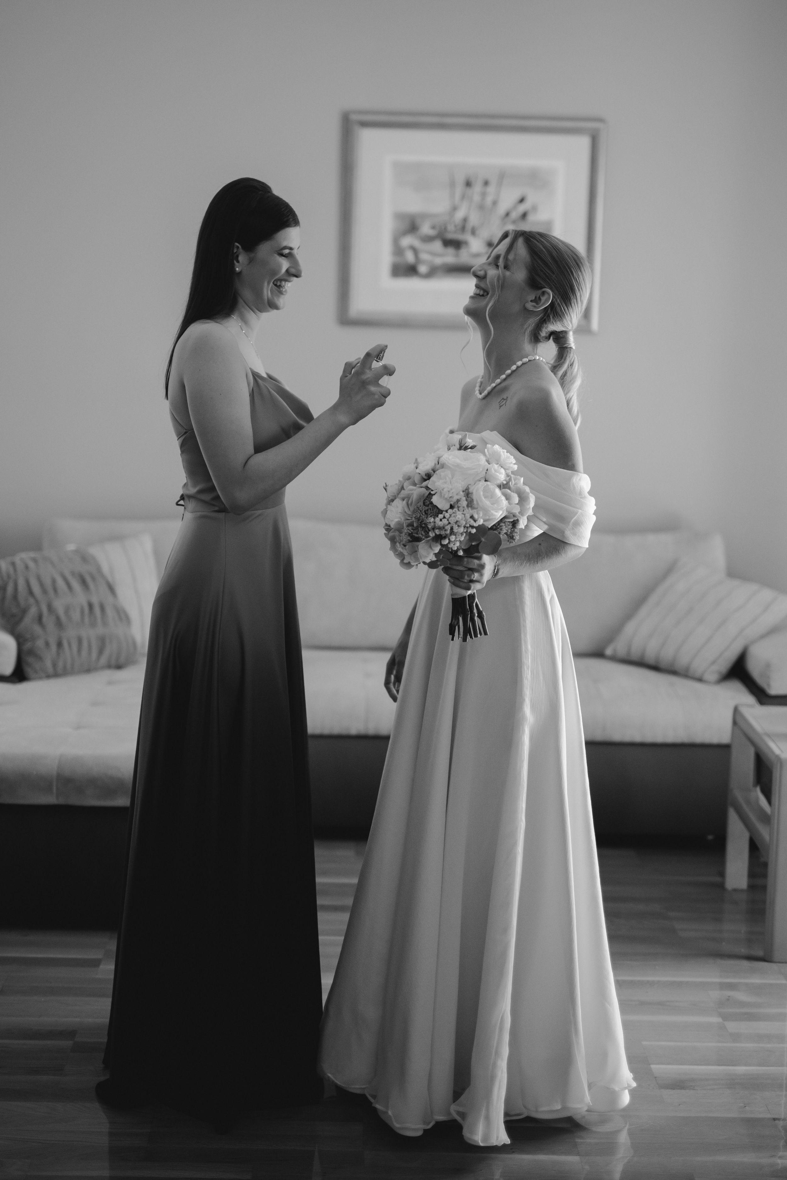 A bridesmaid in a black gown converses with a bride holding a bouquet in a bright interior setting.