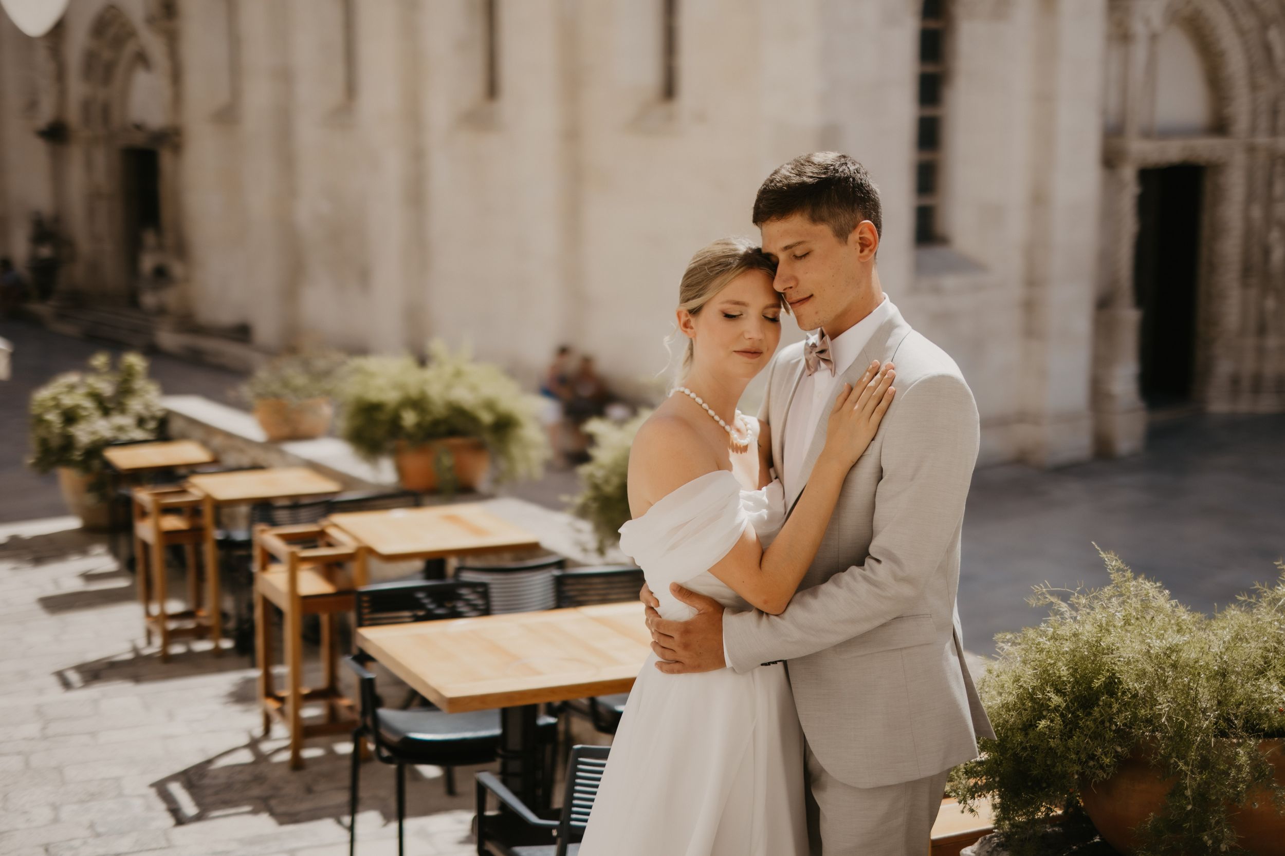 A romantic wedding couple embraces outside a European cafe with outdoor seating.