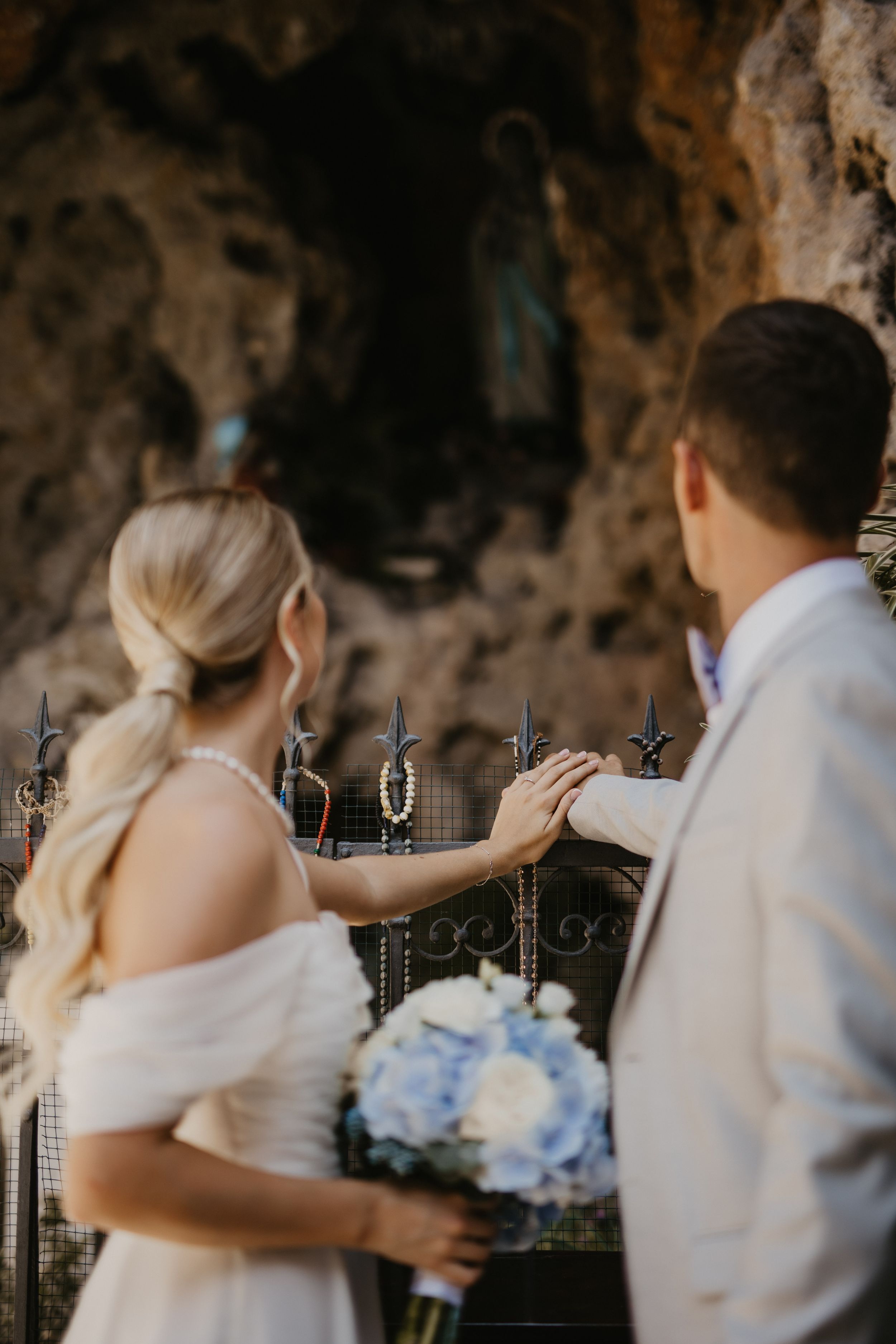 A wedding couple stands before a grotto holding a blue and white bouquet.