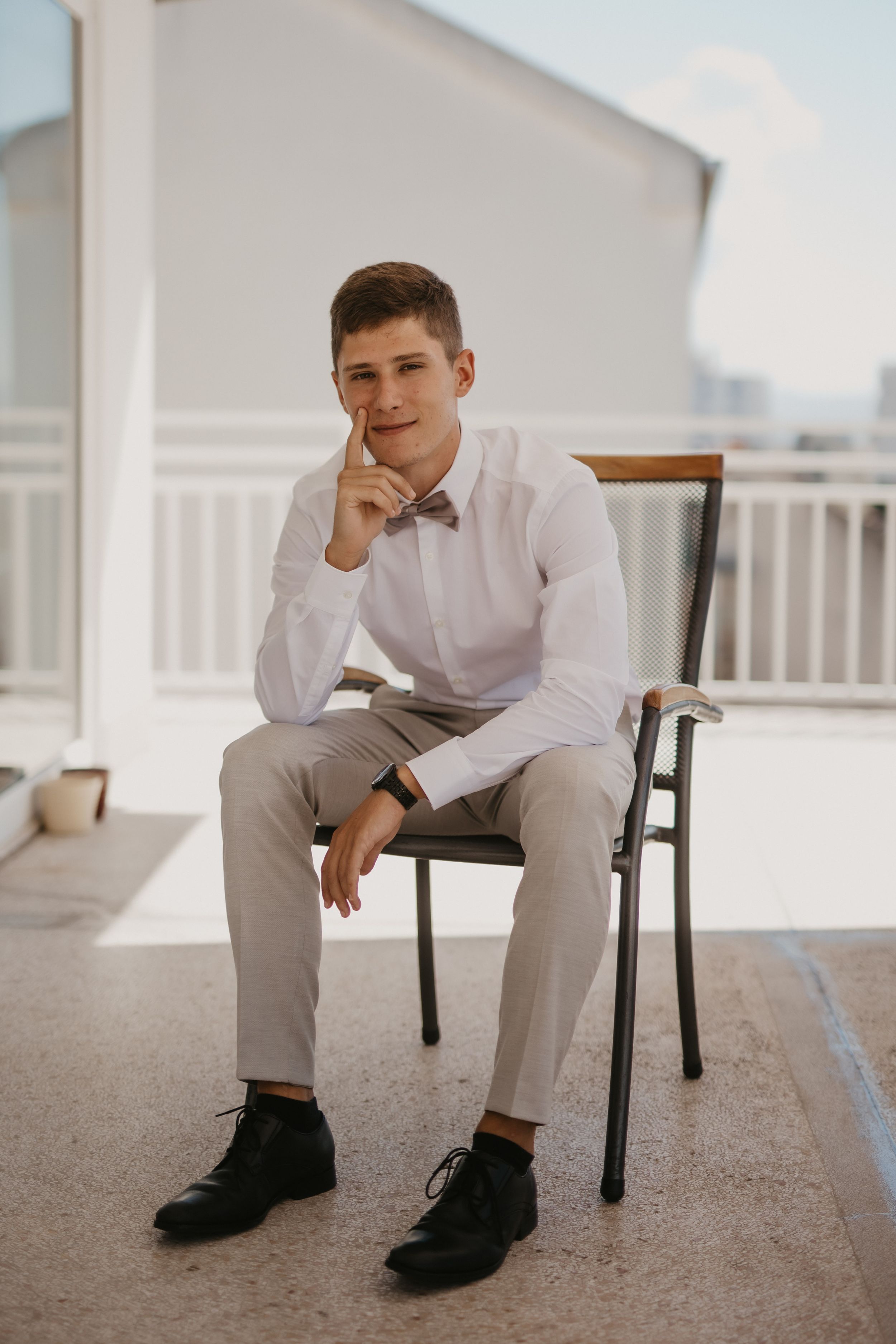 Person in formal white shirt and bowtie sits thoughtfully on outdoor balcony.