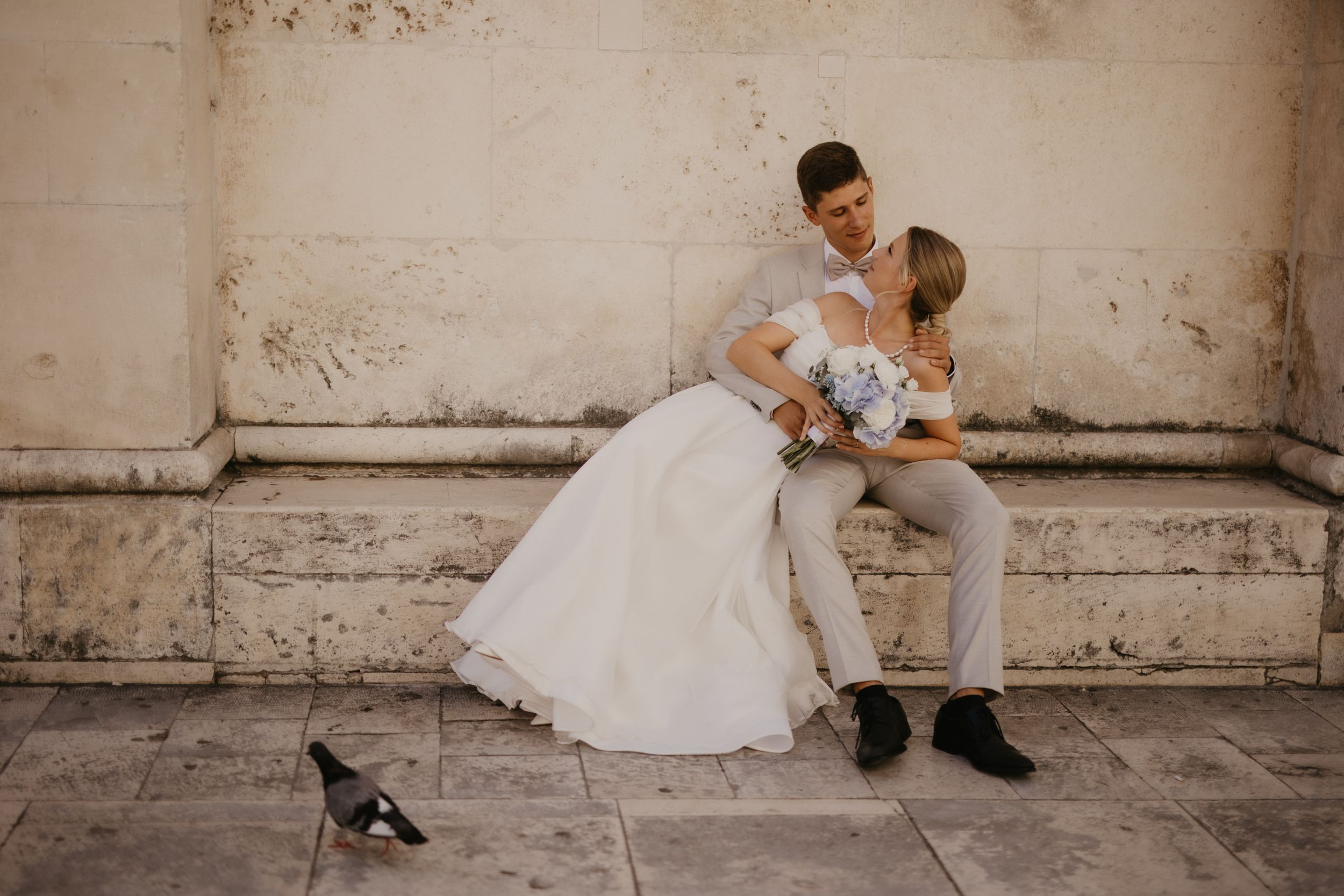 Wedding couple shares intimate moment against weathered wall with cat walking by.