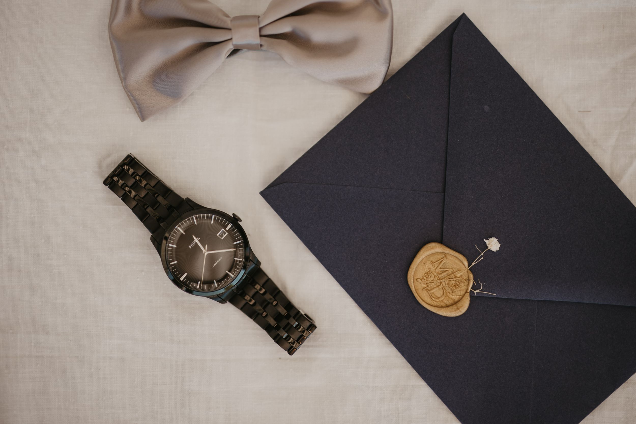 Wedding accessories including beige bowtie, watch and sealed envelope with wax stamp.