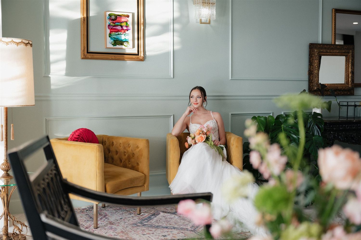 A bride in a white wedding gown sits elegantly in a mustard yellow velvet chair with floral arrangements in the foreground.