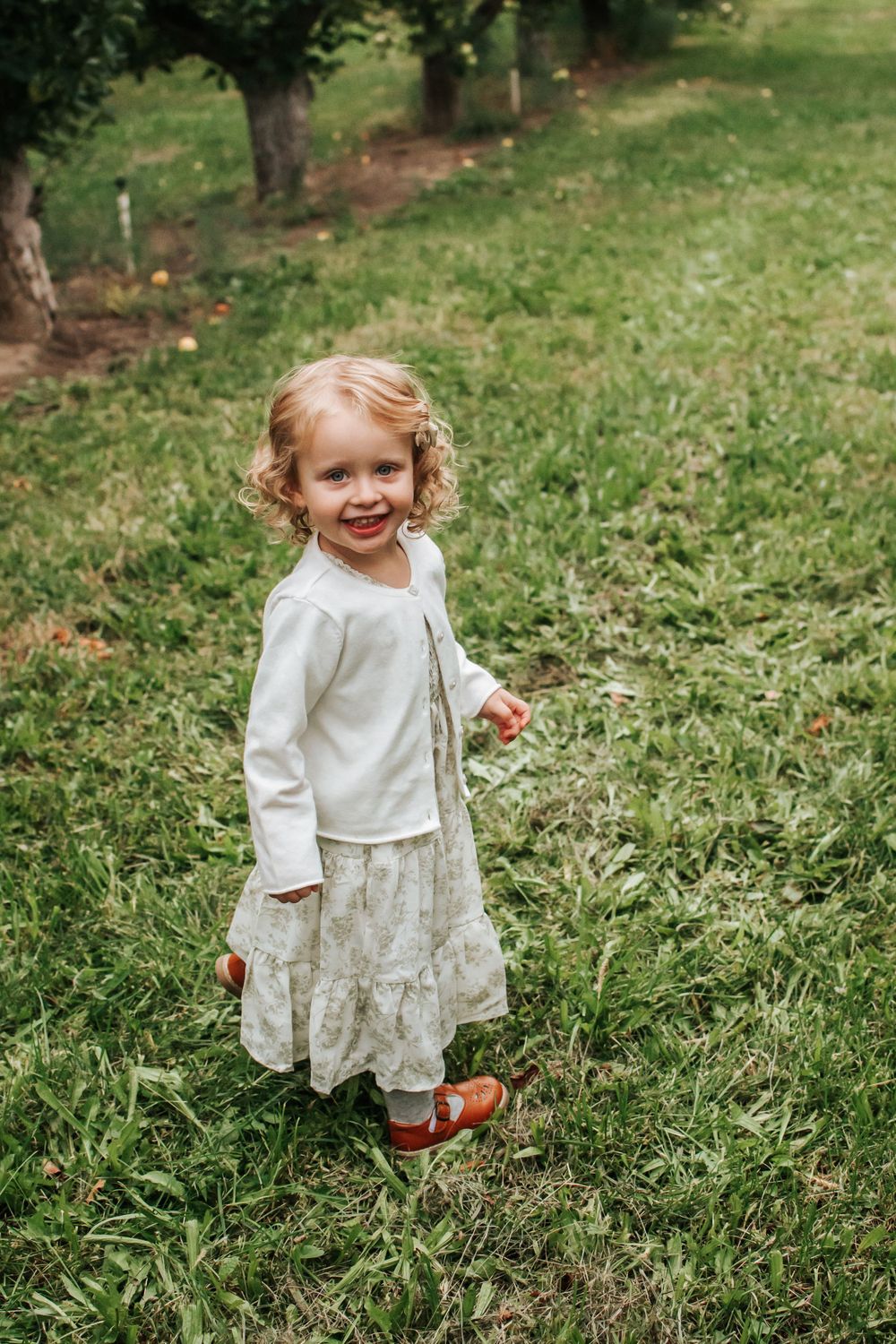 Young child in white sweater and floral skirt smiling while standing in an apple orchard in Benton County, Washington.