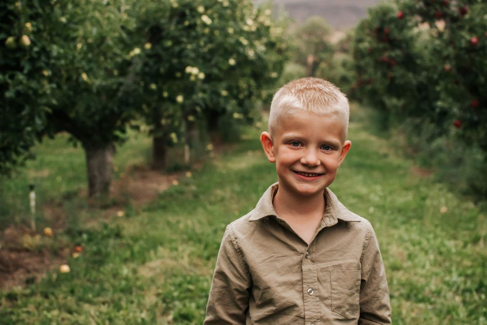 Smiling child wearing brown shirt standing in an apple orchard with green trees and hills in background in Benton City, Washington.