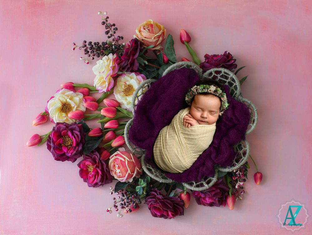Newborn baby wrapped in white fabric nestled in purple floral arrangement with pink roses on pink background.