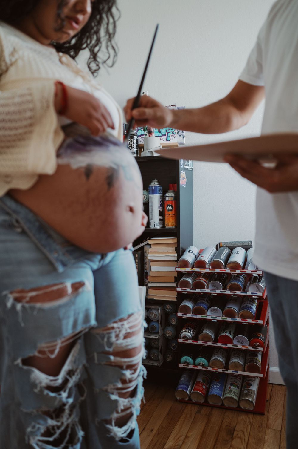 A pregnant figure in distressed jeans stands next to a shelf displaying rows of colorful beverages.