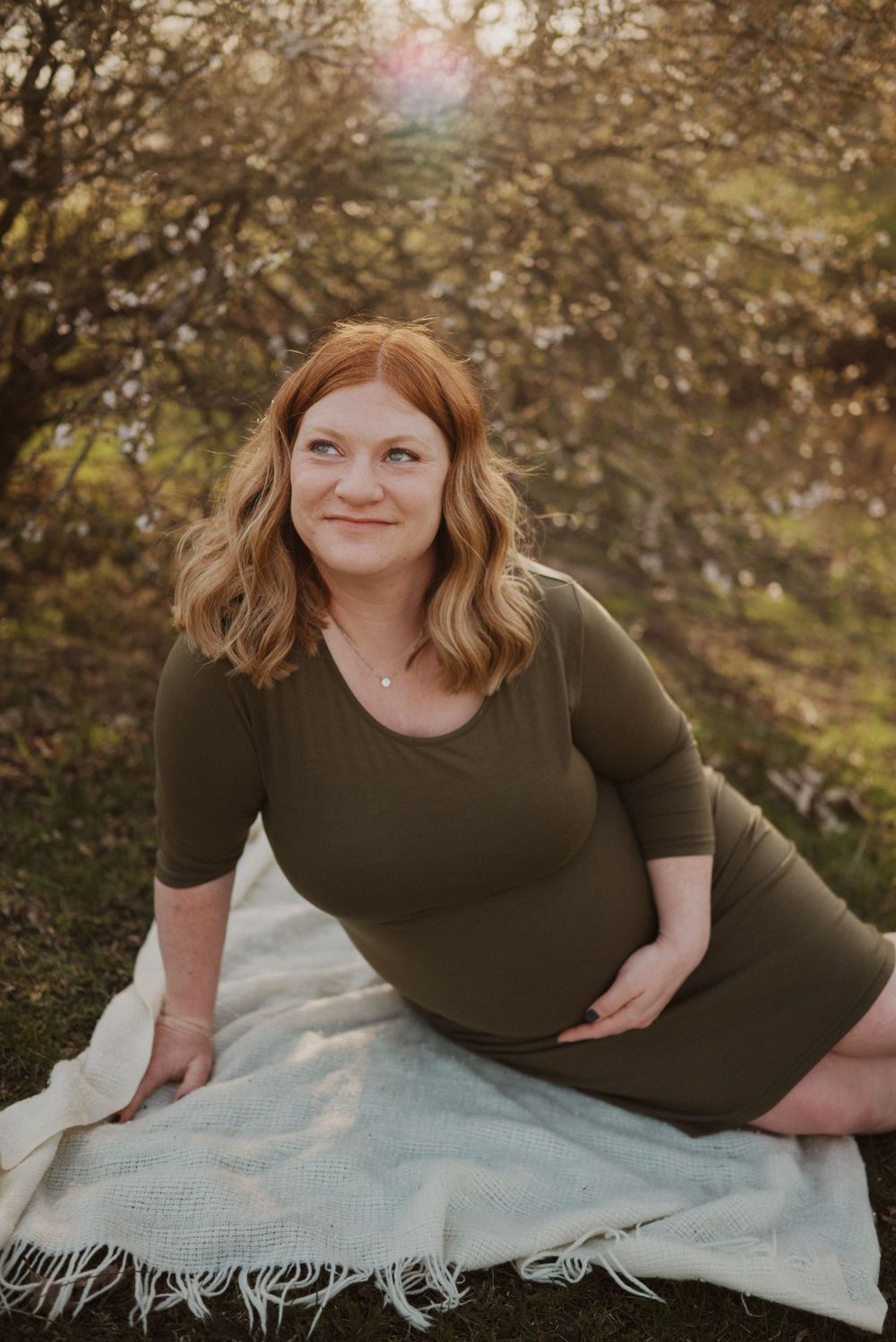A maternity portrait of someone in an olive green dress sitting on a blanket outdoors.