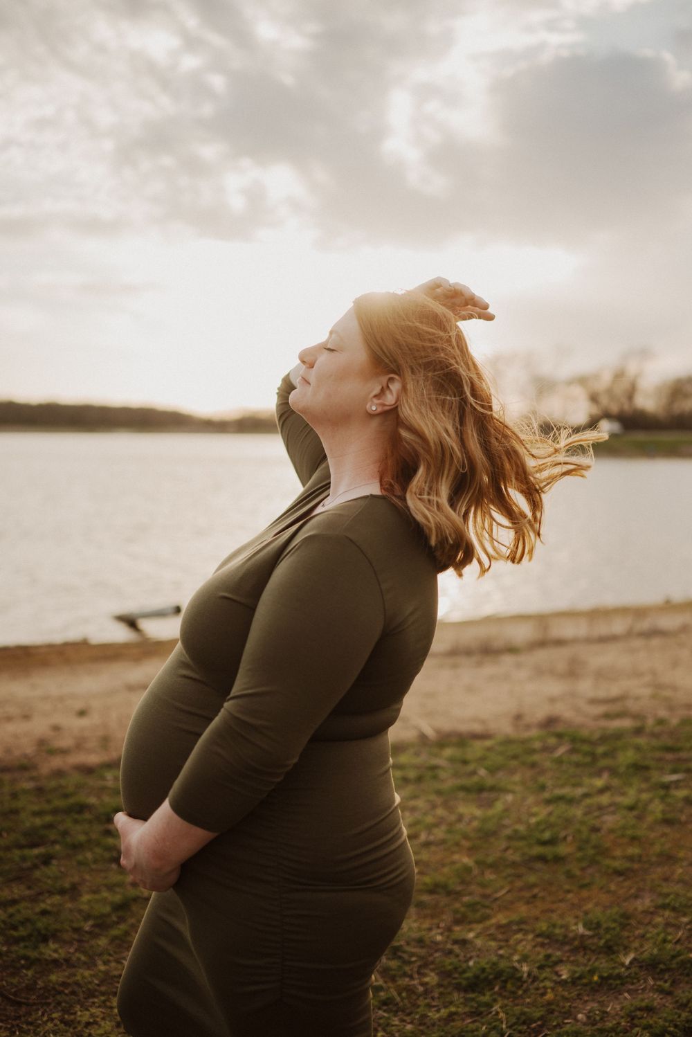 A silhouetted maternity portrait at sunset near a lake with wind-blown hair.