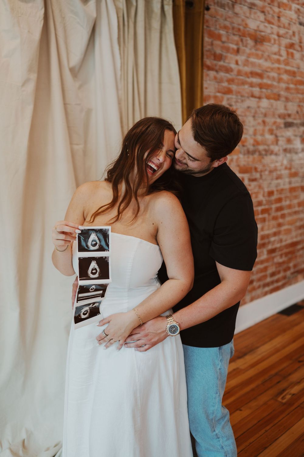 Couple sharing intimate moment while holding ultrasound photos against brick wall.