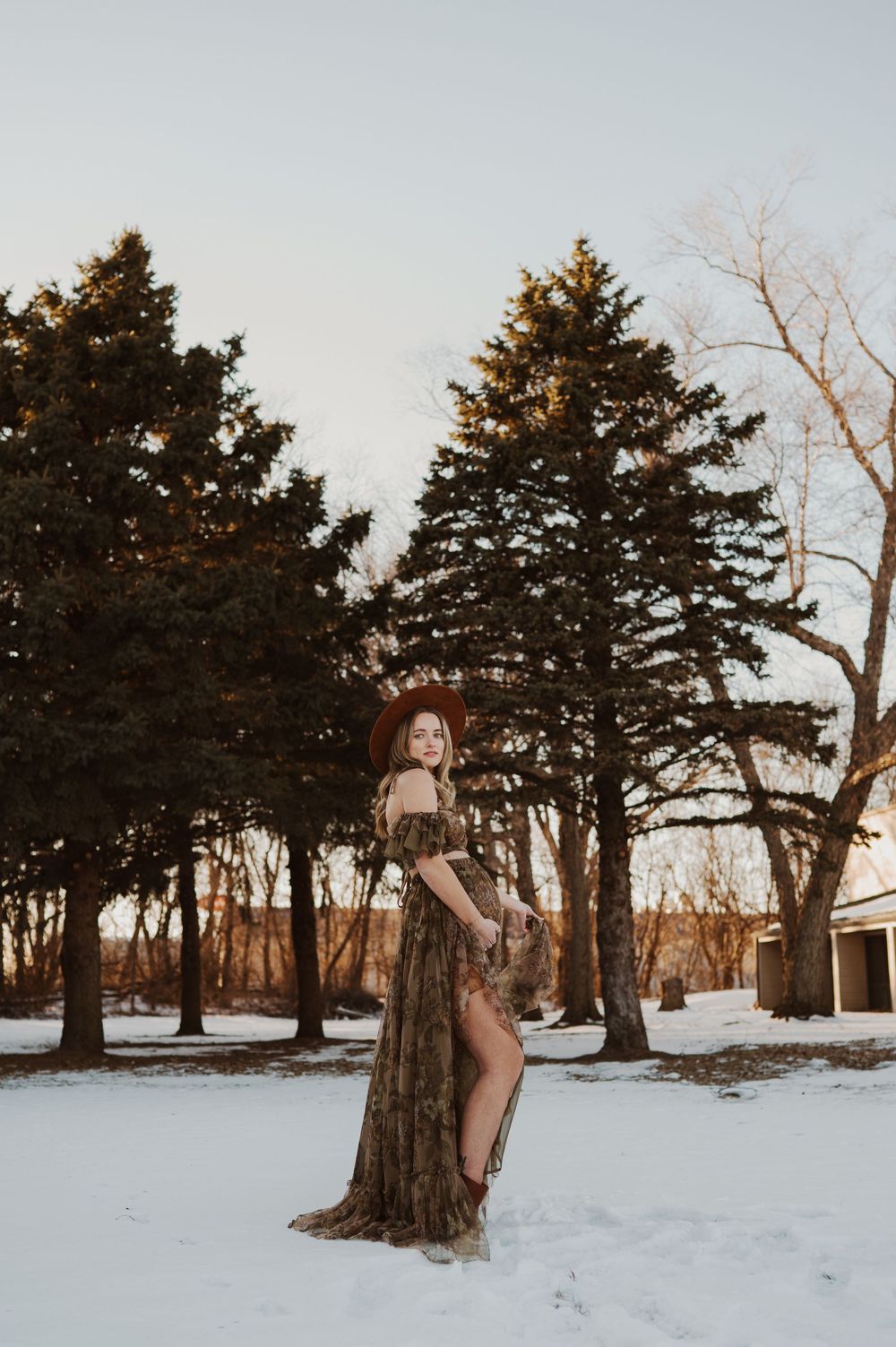 Woman in camo gown and hat poses gracefully in snowy landscape with evergreen trees at golden hour.