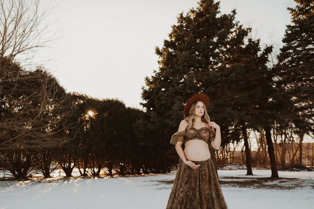 Pregnant woman in floral gown and hat poses outdoors in snowy park with evergreen trees at golden hour.