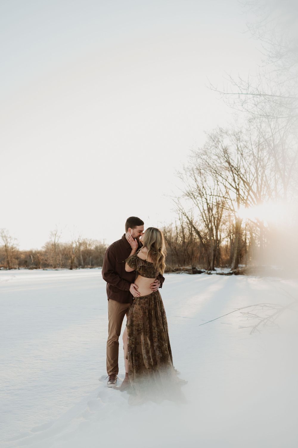 A couple shares a romantic kiss in a snowy winter landscape at golden hour, woman in floral gown, man in burgundy shirt.