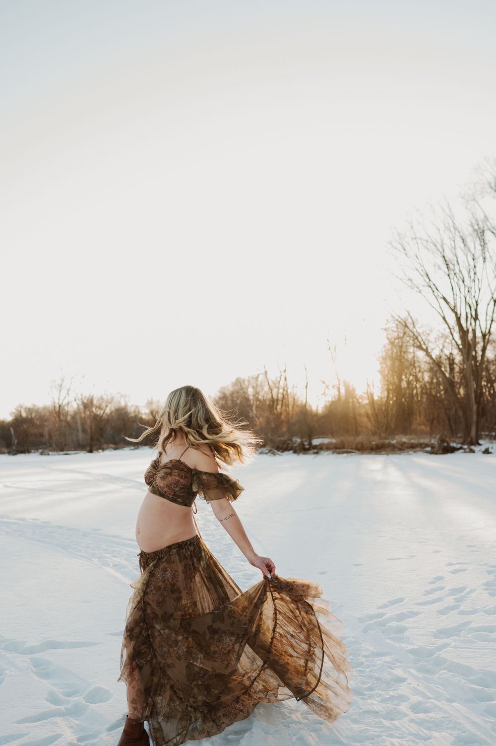 Pregnant woman in brown boho dress twirling in snowy winter landscape at golden hour sunset.