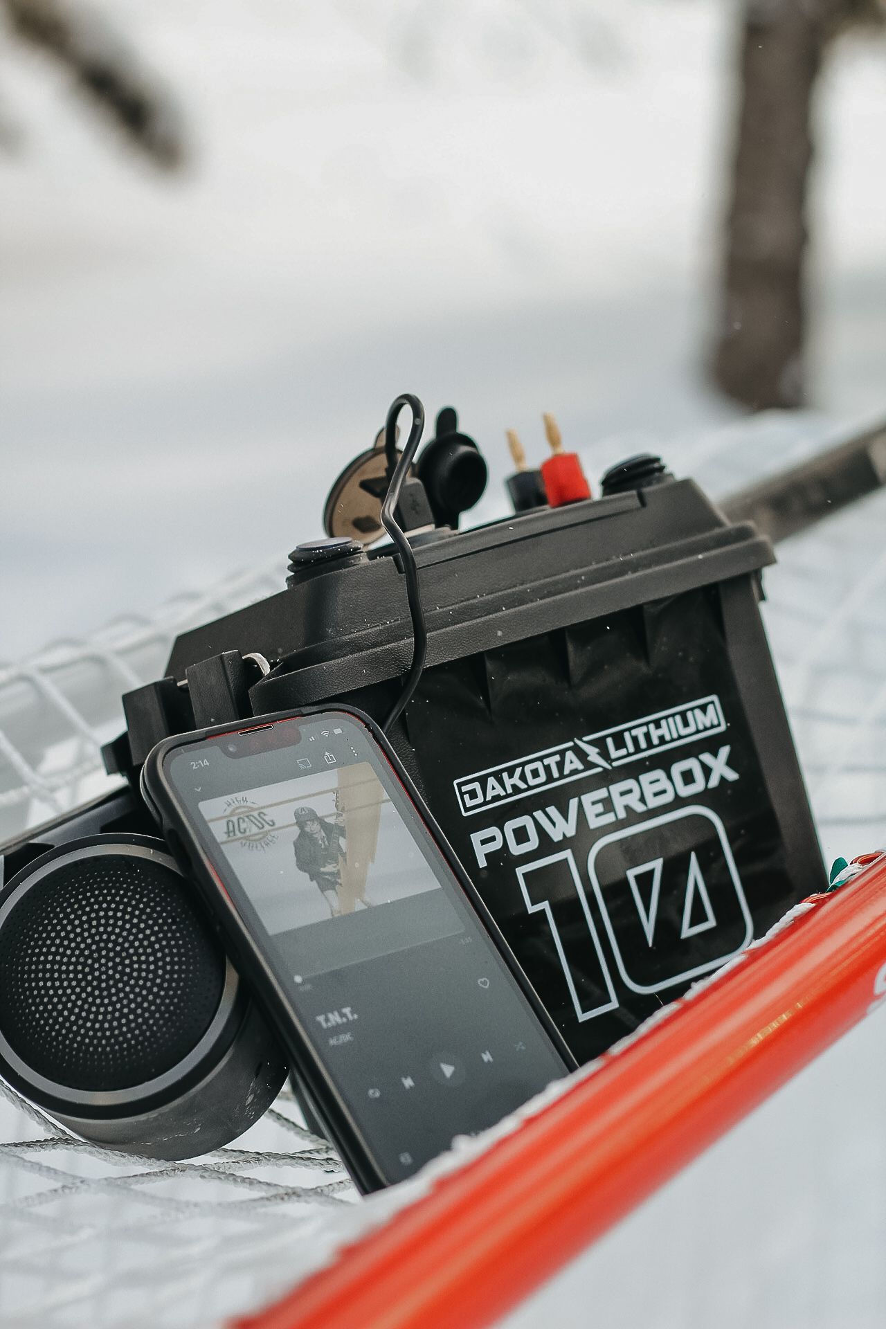 Dakota Lithium power bank with speaker and phone displaying music interface on snowy surface.