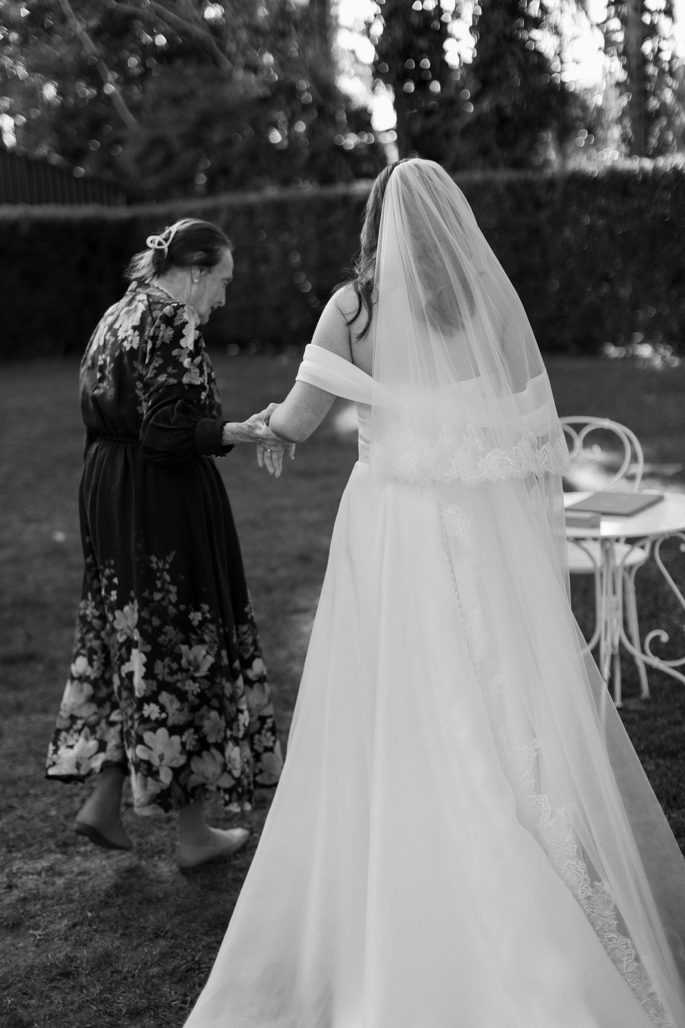 A black and white wedding photo showing final dress adjustments being made outdoors in a garden setting.