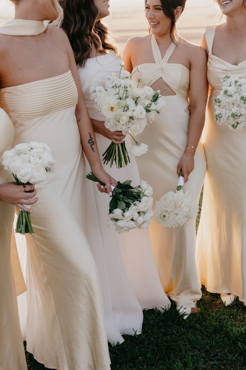 Bridesmaids in elegant cream-colored dresses hold white floral bouquets while standing in a row outdoors.