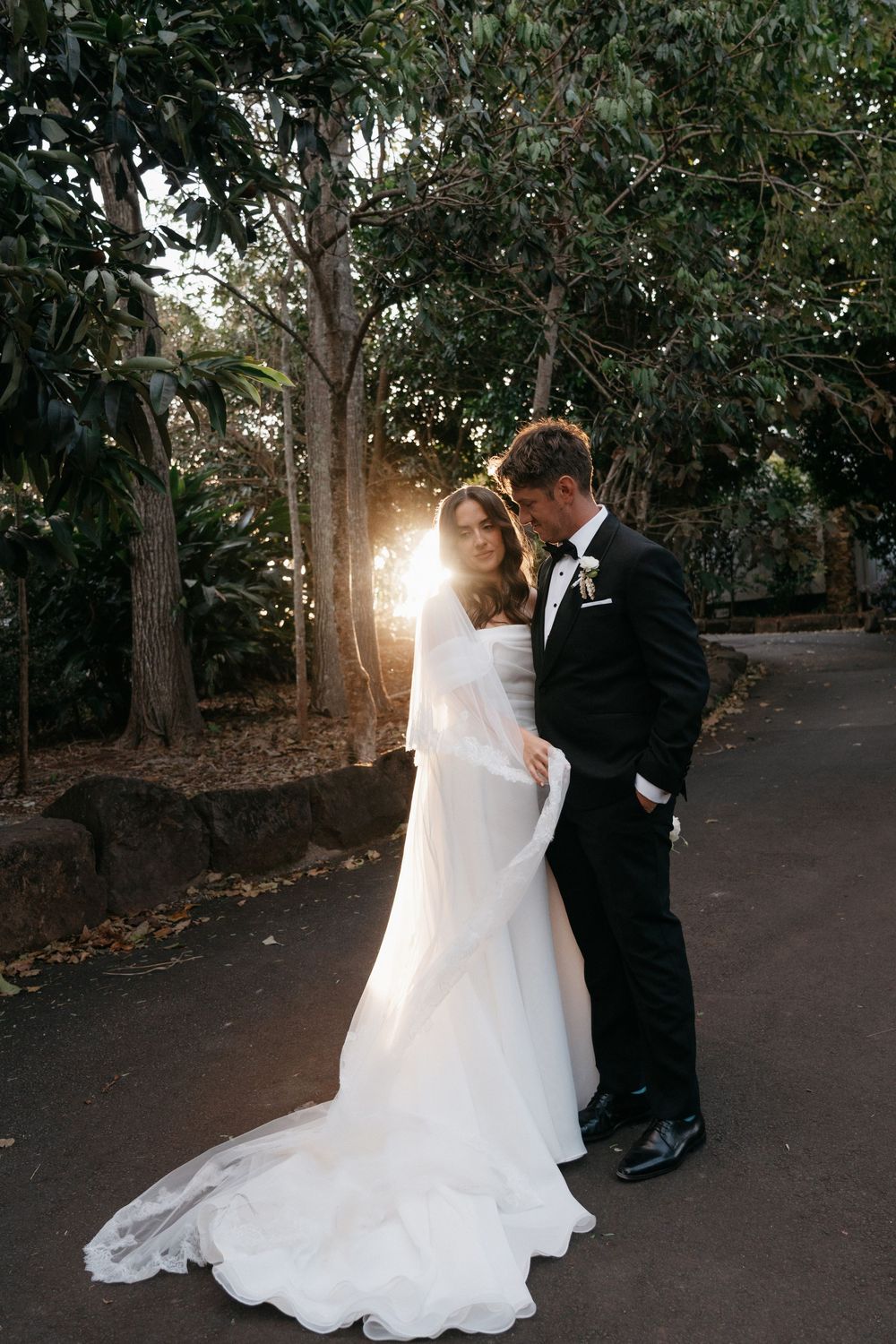 A couple in wedding attire dance together on a tree-lined path at sunset, creating a romantic silhouette.