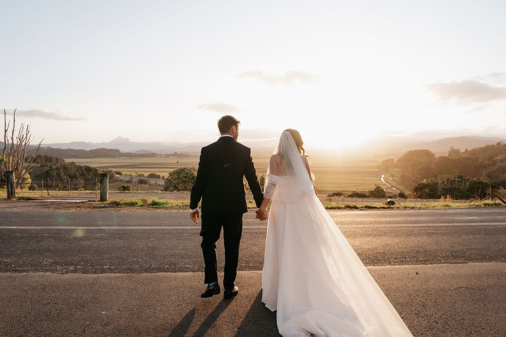 A couple in wedding attire walks through a desert landscape at sunset with mountains in the background.
