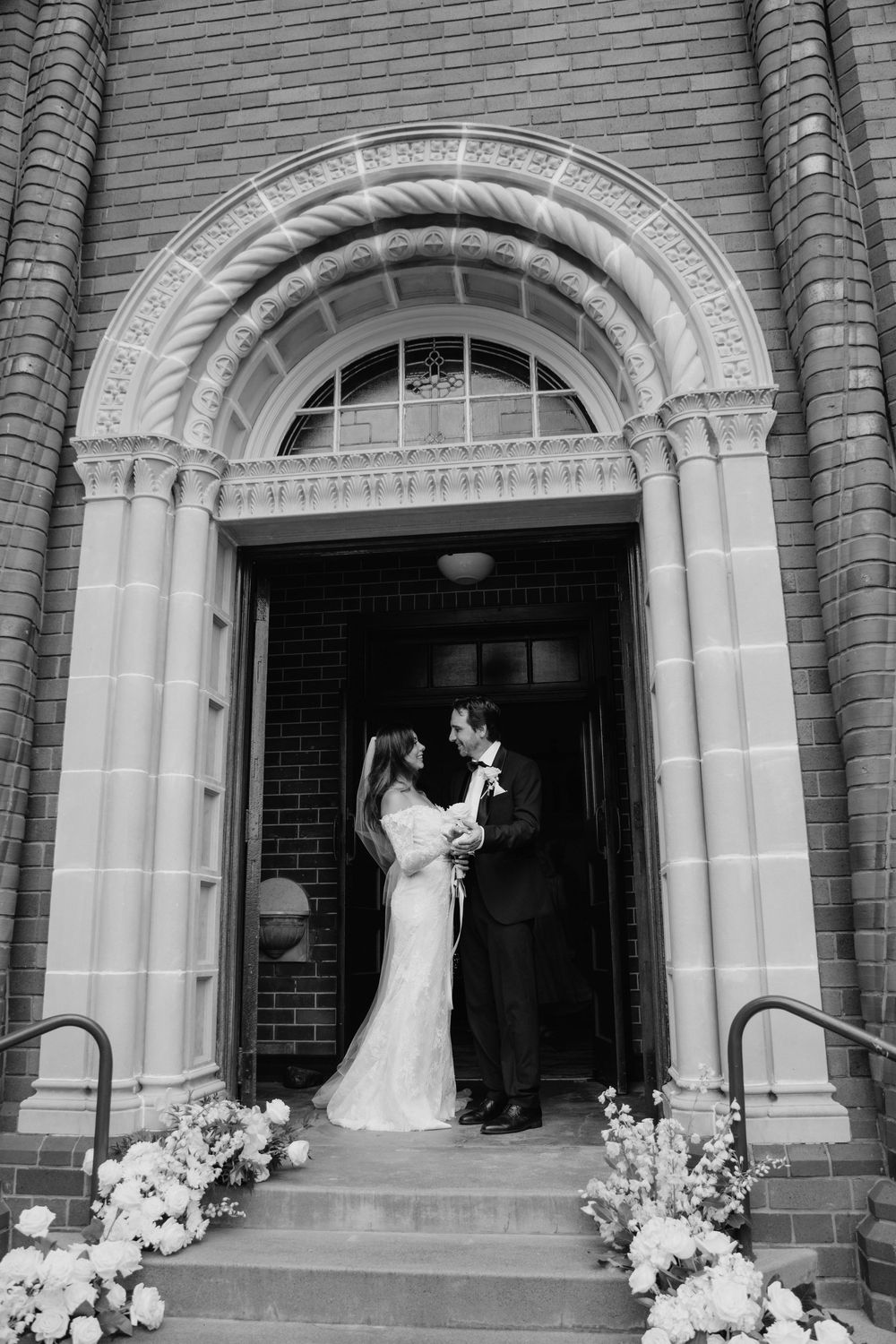 Black and white photo of a wedding couple entering different archways of a historic brick building decorated with flowers.