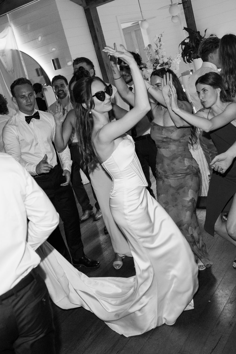 Bride in white gown and sunglasses dancing joyfully with guests at a lively wedding reception in black and white.
