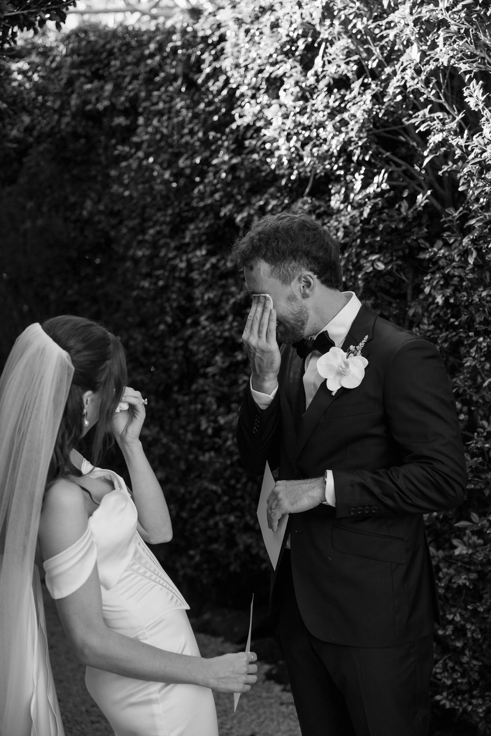 Black and white photo of emotional bride and groom crying together on wedding day near lush green hedge backdrop.