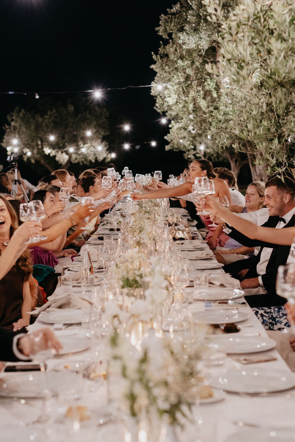 Wedding guests raise glasses in a toast at an elegant outdoor reception table decorated with string lights and floral centerpieces.