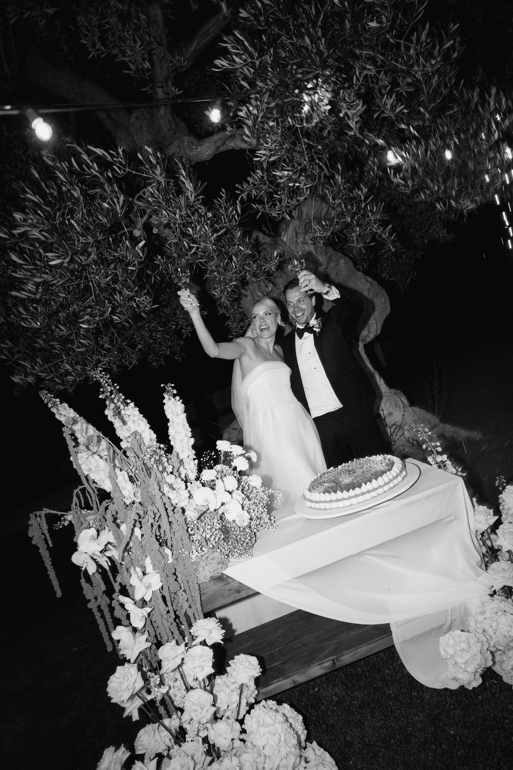 A wedding celebration captured in black and white as people dance joyfully in an outdoor garden setting with decorative lights.