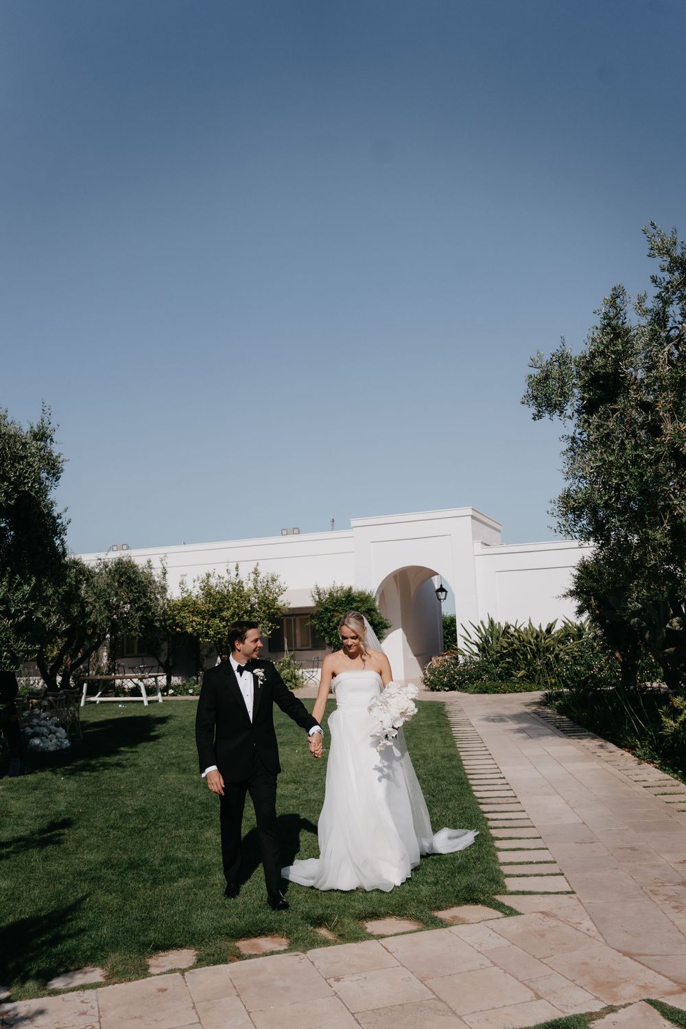 Newlyweds share a romantic moment walking together on a sunny day along a concrete path surrounded by green grass.