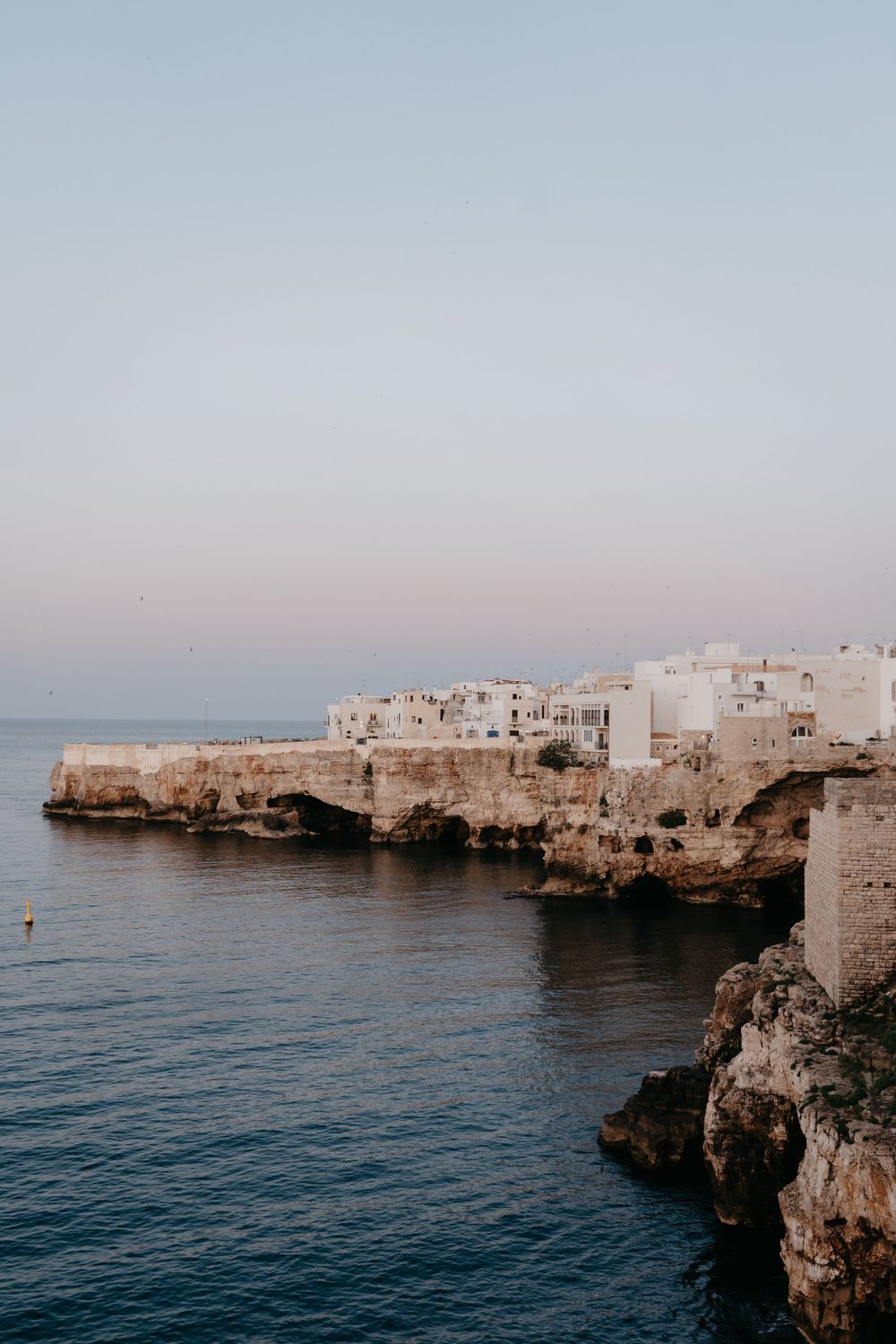 White stone buildings perch dramatically on limestone cliffs overlooking the deep blue Mediterranean Sea at sunset.