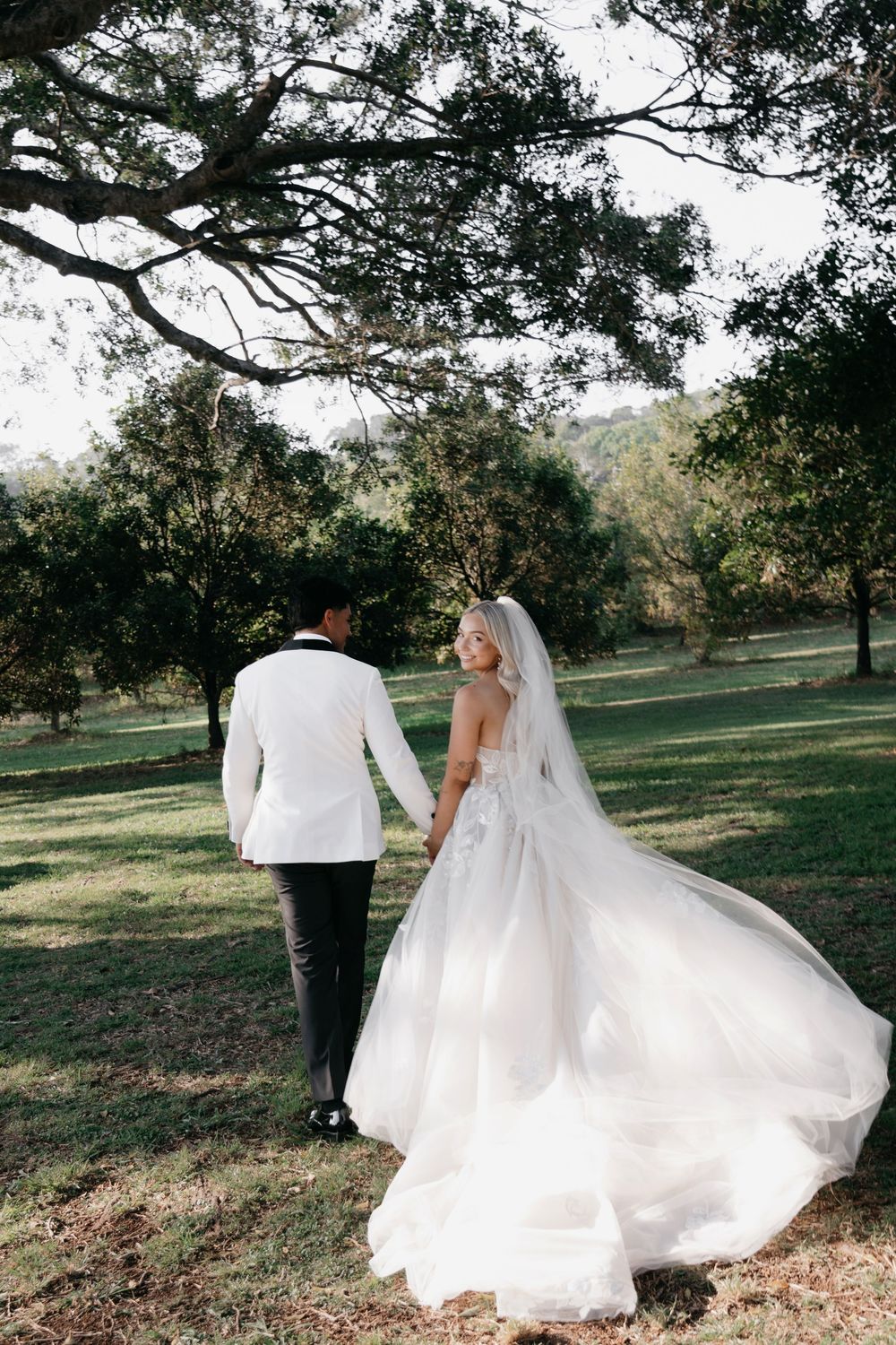 A couple in wedding attire walks through an outdoor setting with sunlit trees and scattered autumn leaves.