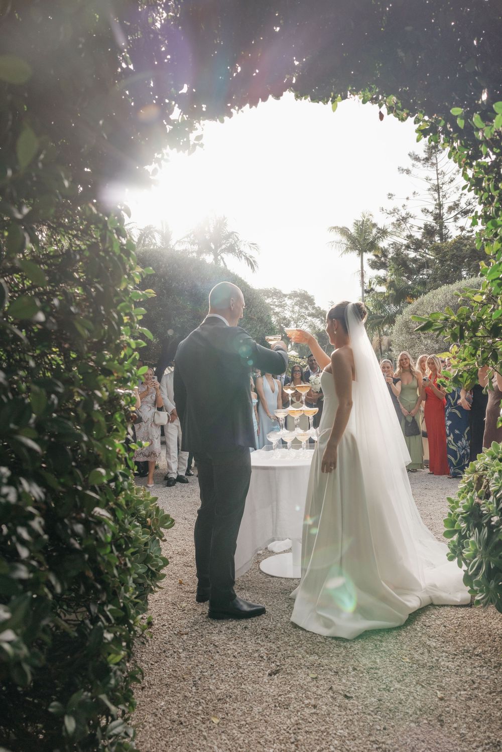 Bride and groom toast champagne under a lush garden arch at an outdoor wedding reception in golden sunlight.