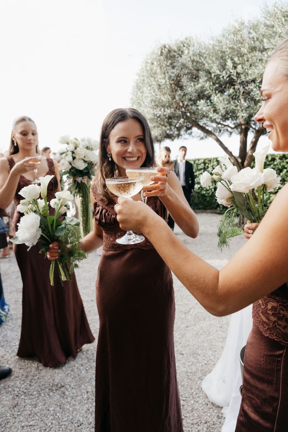 Bridesmaids in brown velvet dresses toasting with champagne coupes while holding white floral bouquets at a wedding.