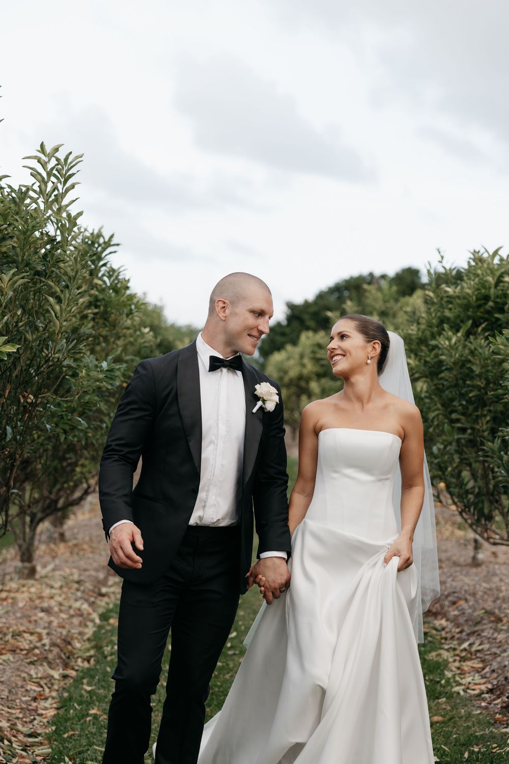 Bride and groom walk hand in hand through lush orchard, smiling at each other in elegant wedding attire.
