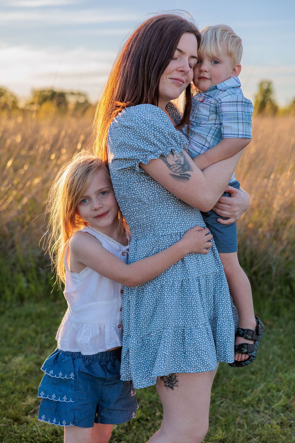 A mother holds her young son while her daughter hugs her in a golden sunlit field at sunset.