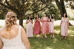 A group of bridesmaids in pink floral and solid dresses react with excitement in a sunny garden setting.