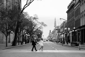 Black and white photo of a couple crossing a wide city street lined with trees and historic buildings.