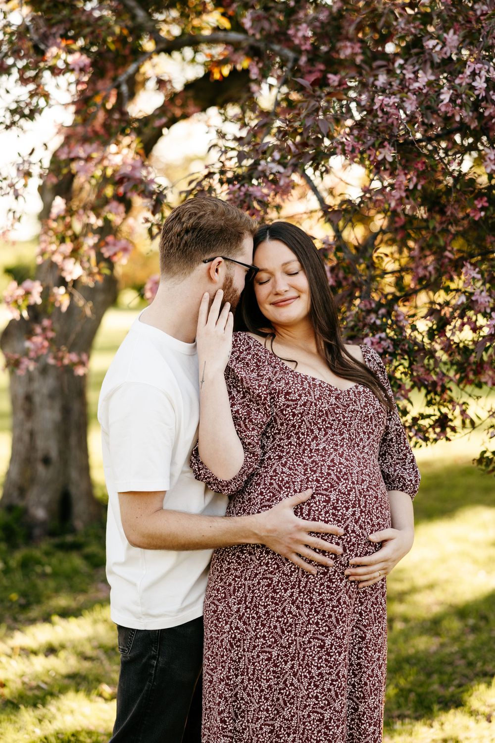 Couple sharing a tender moment under blooming pink tree, pregnant woman in floral dress cradling baby bump outdoors.