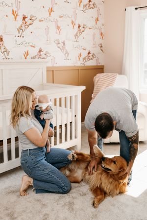 Young family with newborn baby and golden retriever dog in a decorated nursery room with floral wallpaper.