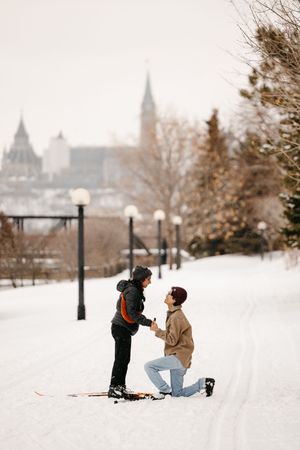Person proposing on one knee in snowy winter park with historic Parliament buildings visible in background.