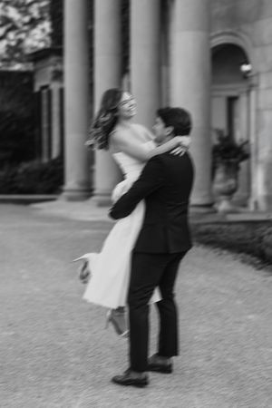 Black and white photo of a groom lifting his bride outdoors near elegant columns at a wedding venue.